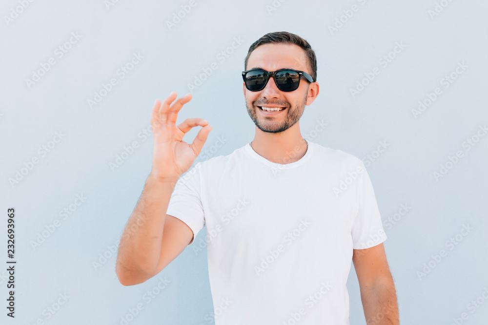 Naklejka premium Young smiling man in sunglasses having happy look, gesturing, showing OK sign or showing okay gesture with his fingers on blue background
