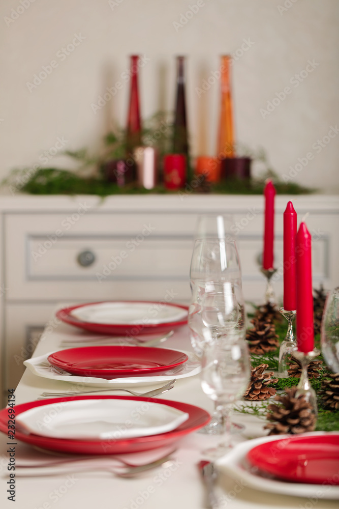 Merry Christmas and Happy New Year! Тable setting festive decor - white and red dishes, red candles in the candlesticks, juniper branches and fir cones. Merry Christmas and Happy New Year! Тable setting festive decor - white and red dishes, red candles in the candlesticks, juniper branches and fir cones.