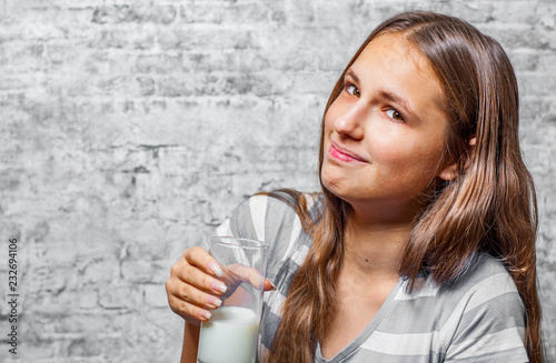 portrait of young teenager brunette girl with long hair holding a glass of milk on gray wall background