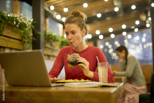 Hungry girl eating cheeseburger and french fries in college cafe while searching for information in the net