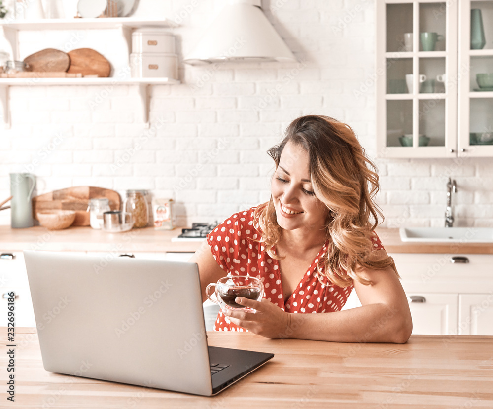 Happy beautiful woman drinking coffee. Woman working on a laptop in kitchen. Happy beautiful woman drinking coffee. Woman working on a laptop in kitchen.