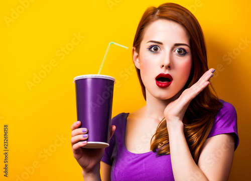 surprsied redhead girl in purple dress with drink on yellow background. photo