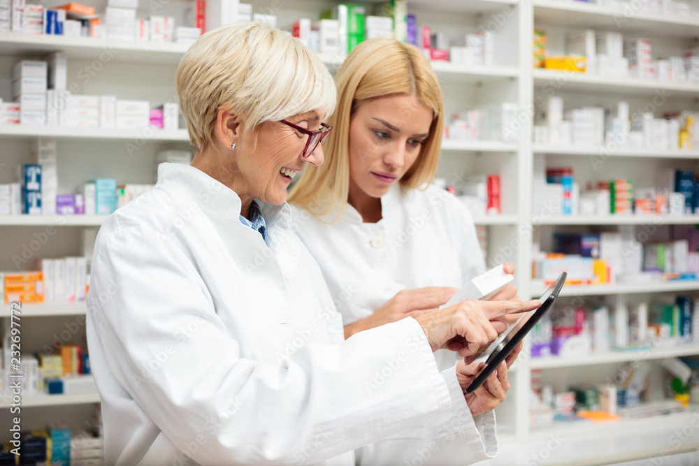 Young and mature female pharmacists standing in front of a shelf with medications. Senior woman using a tablet and showing something to her younger colleague. Pharmaceutics and healthcare concept