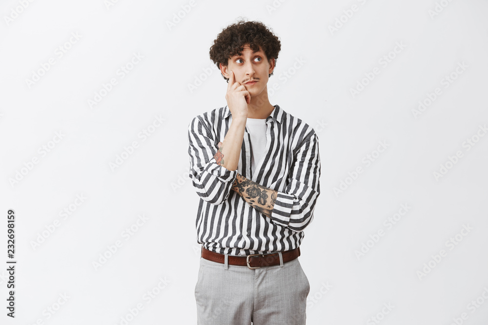 Curious thoughtful and cute jewish guy with dark curly hair and moustache holding hand on face while thinking looking at upper right corner curiously seeing interesting thing over gray wall Curious thoughtful and cute jewish guy with dark curly hair and moustache holding hand on face while thinking looking at upper right corner curiously seeing interesting thing over gray wall