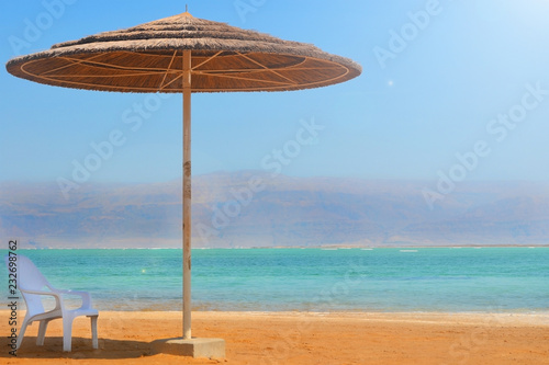Fototapeta Naklejka Na Ścianę i Meble -  A straw umbrella and a white chair stand on a sandy beach near the water. Rest on the Dead Sea in Israel overlooking the mountains of Jordan