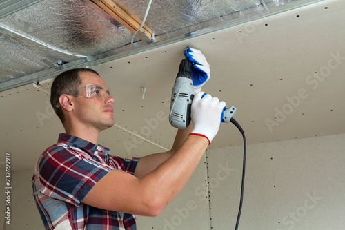 Young man in goggles fixing drywall suspended ceiling to metal frame using electrical screwdriver on ceiling insulated with shiny aluminum foil. Renovation, construction, do it yourself concept. photo