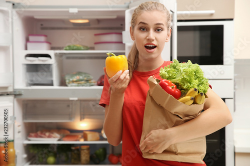Beautiful young woman with paper bag full of food near refrigerator at home
