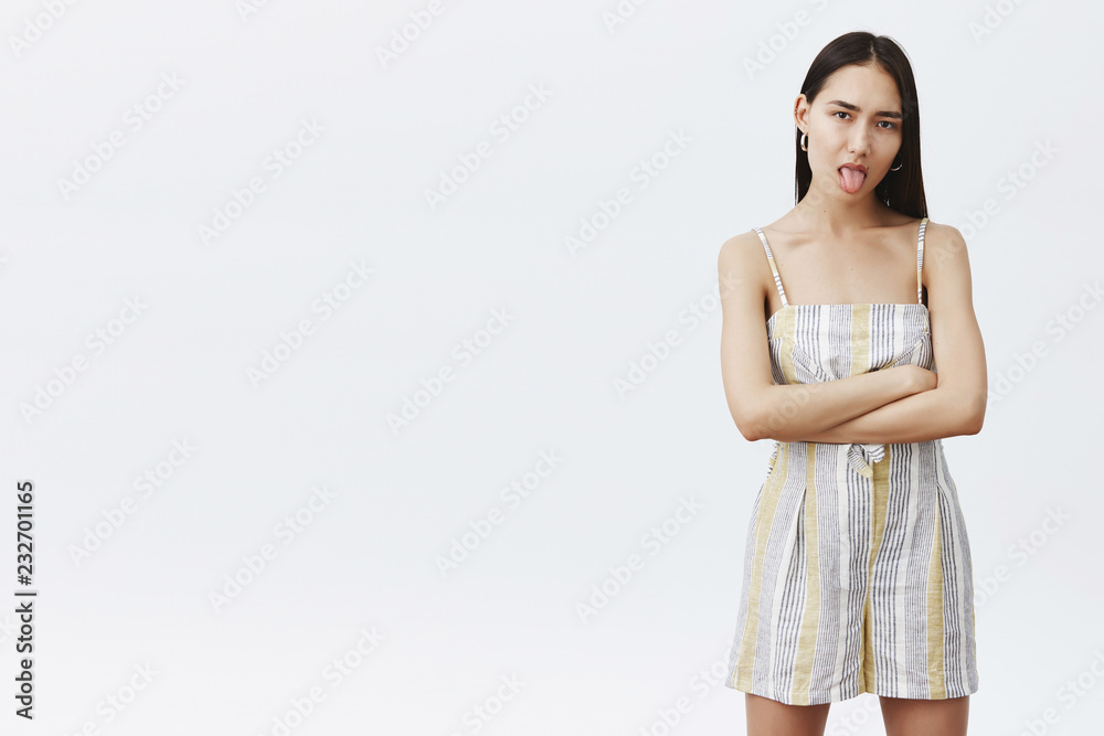 Indoor shot of moody good-looking fashionable woman in matching outfit, holding hands crossed in confident pose, sticking out tongue and staring moody and displeased at camera over grey wall Indoor shot of moody good-looking fashionable woman in matching outfit, holding hands crossed in confident pose, sticking out tongue and staring moody and displeased at camera over grey wall