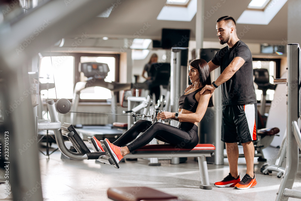 Beautiful slim girl dressed in black sport clothes is doing exercises for the abdominals on a special exercise machine under the supervision of a coach in the gym Beautiful slim girl dressed in black sport clothes is doing exercises for the abdominals on a special exercise machine under the supervision of a coach in the gym