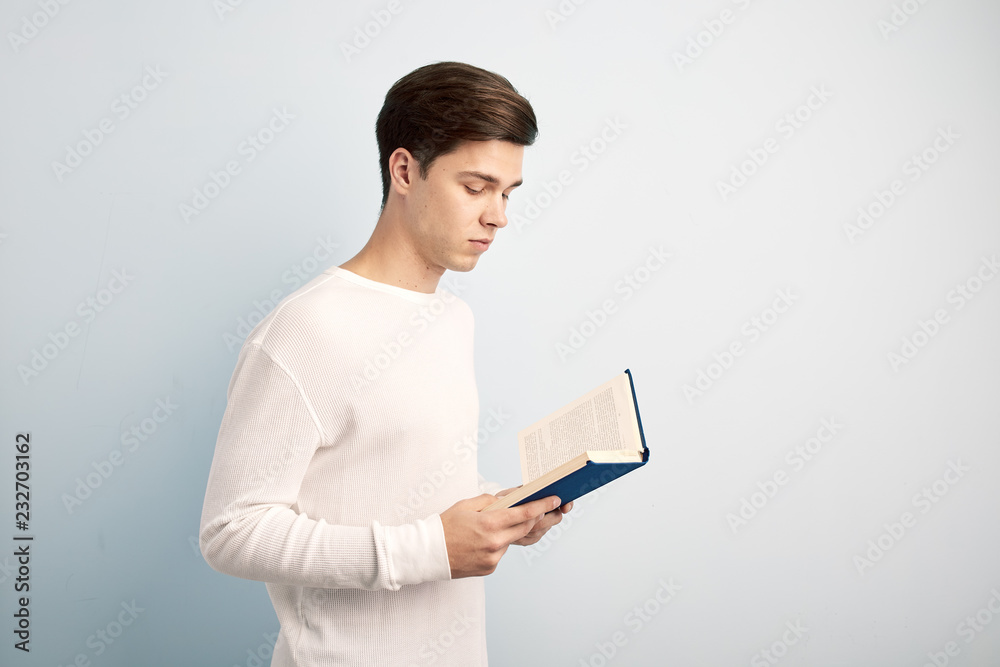 Dark-haired guy dressed in a white long sleeve t-shirt and jeans reads a book in his hand on a white background