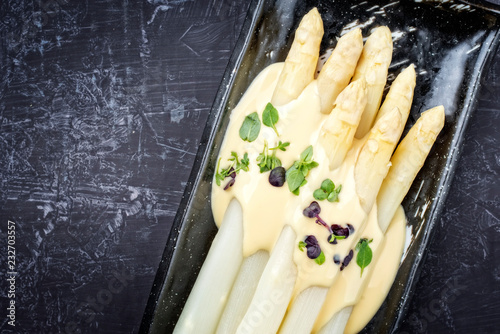 Traditional boiled white asparagus with sauce hollandaise and herbs as top view on a plate with copy space left