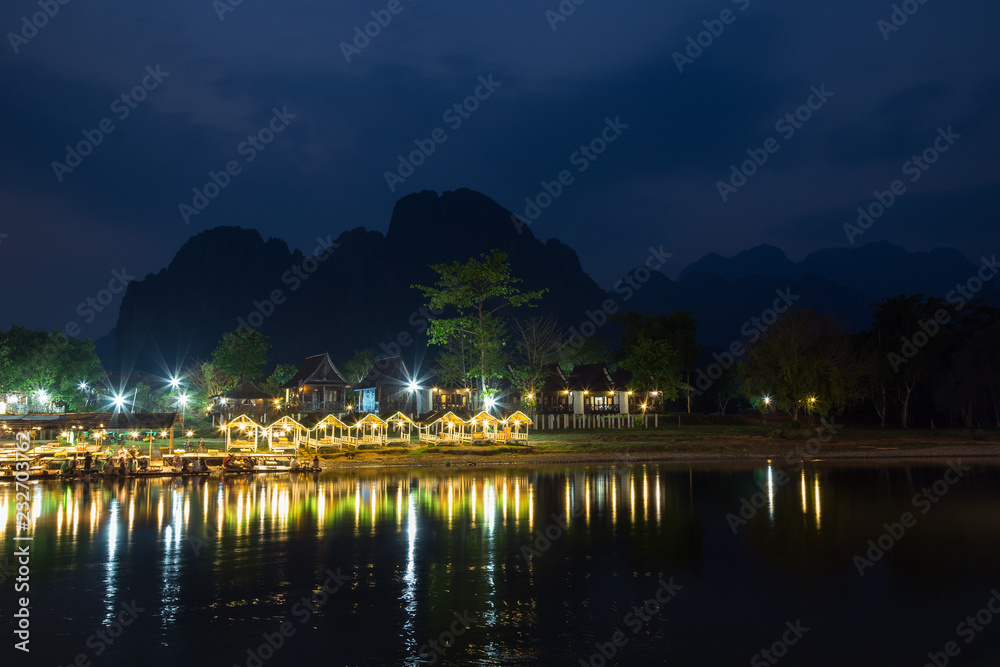 Fototapeta premium Several people at a lit waterfront restaurant by the the Nam Song River and silhouette of karst limestone mountains in Vang Vieng, Vientiane Province, Laos, at dusk.