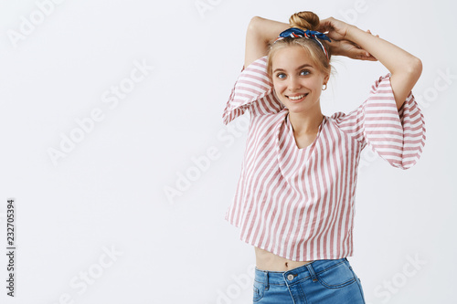 Studio shot of tender and feminine cute blonde female with combed hair and striped blouse raising hands behind head and posing in modeling pose over gray background smiling at camera