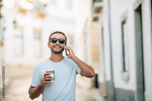 Handsome young man dressed in trendy wear strolling on city street having phone conversation using international operator and roaming holding coffee to go © F8  \ Suport Ukraine