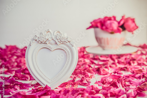 Heart shaped vintage style blank photoframe on the white table with fresh petals and bouquet of pink purple peonies in cup. Love, romantic, Valentines day concept. Selective focus. Copy space.