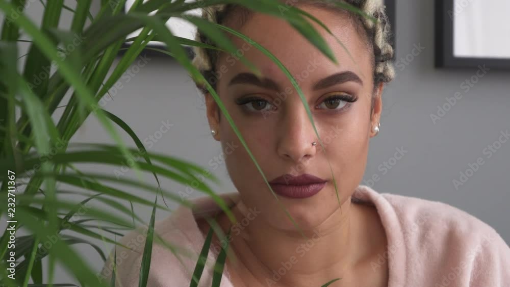 Beautiful serious young woman looking into camera near houseplant Beautiful serious young woman looking into camera near houseplant