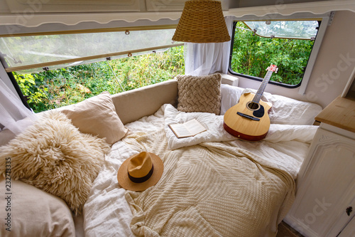 Inside the camper van. Unfilled bed, pillows, guitar, book, hat, white wooden decoration of the house on wheels.