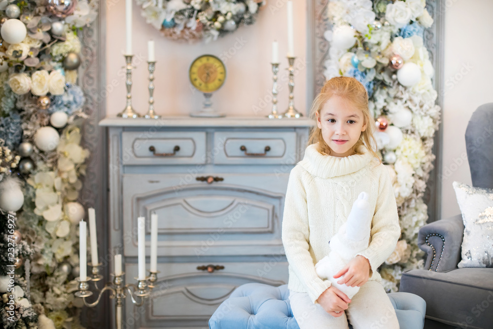 Merry Christmas and Happy New Year! Charming little blonde girl in white warm sweater with toy bear sitting on the blue armchair near Christmas tree Merry Christmas and Happy New Year! Charming little blonde girl in white warm sweater with toy bear sitting on the blue armchair near Christmas tree