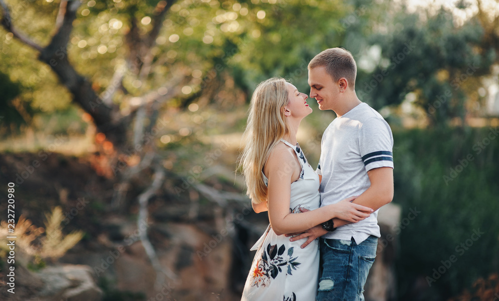 Fototapeta premium Stylish guy and a young girl hugging against the backdrop of nature and sunset with backlight. Portrait of beautiful people.