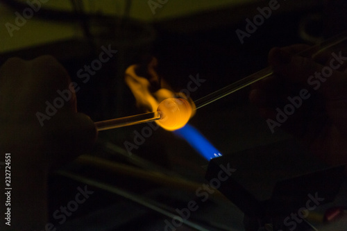 A crafts person is using a blow torch to melt and form a piece of glass. The flame is blue and golden where it is hitting the glass. 