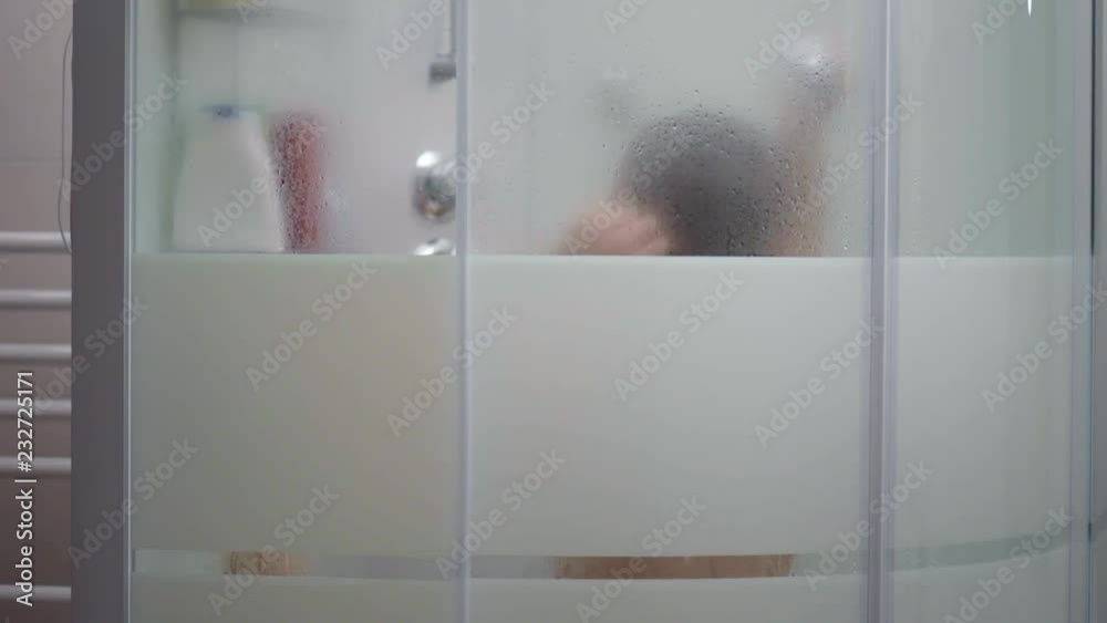 Young girl bathing under a shower at home - behind glass. Beautiful teen girl bathing under a shower in the bathroom. Happy child washing head, face and body with water. Stock ビデオ | Adobe Stock