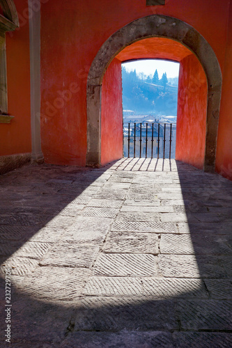 Bright winter sunlight casts a dramatic shadow as it shines through a window cut out of a red painted wall, overlooking the town of Castelnuovo di Garfagnana in the province of Lucca in Tuscany, Italy