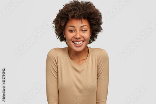 Smiling dark skinned woman has Afro hairstyle, shows white perfect teeth, glad to recieve good mark on exam, wears beige sweater, models against studio background. Ethnicity and emotions concept