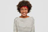 Happiness concept. Beautiful black woman with Afro haircut, smiles gladfully at camera, dressed in striped clothing, models against white background, being in high spirit as rejoices promotion at work