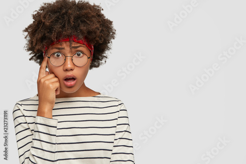 Astonished pleasant looking woman keeps breath from amazement  index finger on temple  shocked to forget something important  wears round spectacles  isolated on white wall  copy space aside
