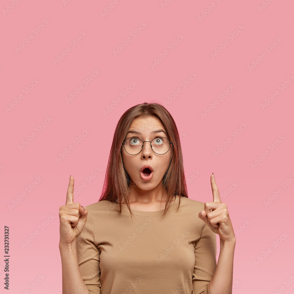 Emotive stupefied European woman points with both index finegrs upwards, shows place for advertisement or text, wears round transparent glasses, poses against pink studio wall. Reaction concept