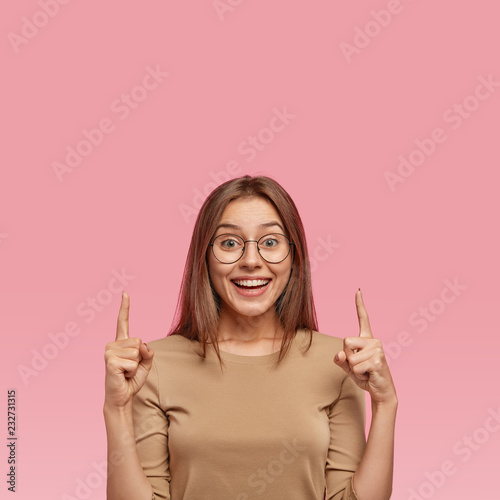 Vertical shot of happy dark haired woman with toothy smile, wears round spectacles, discusses amuzing copy space or place to go, dressed in beige sweater, points at blank copy space upwards.