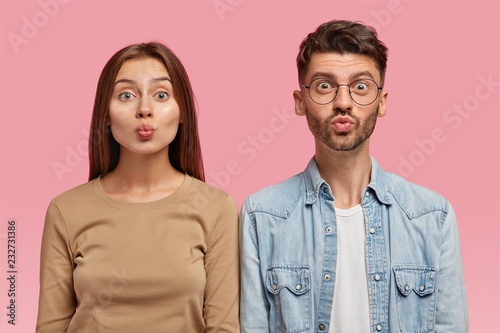 Portrait of attractive girlfriend and boyfriend make grimace, pout lips, have funny looks, dressed casually, stand shoulder to shoulder, model against pink background. Friendship, facial expressions
