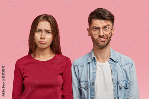 Horizontal shot of gloomy woman and man with puzzled expressions, have displeased sad looks, stand closely to each other, pose against pink background. Negative feelings and emotions concept