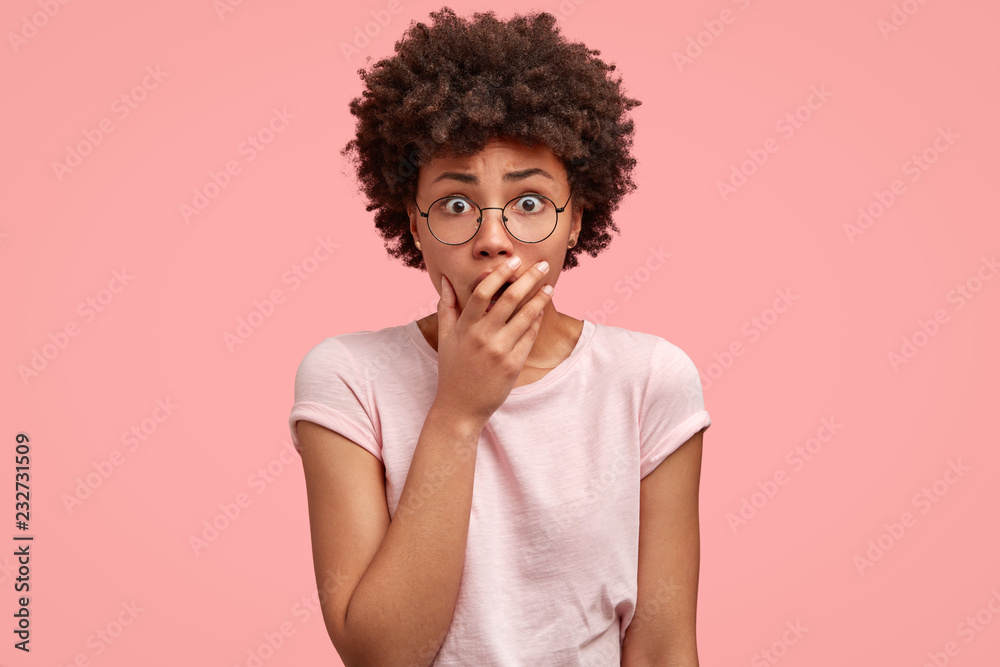 Emotive fearful housewife keeps hand on mouth, gazes at camera with eyes full of surprisement and disbelief, wears glasses, models against pink studio wall. Mute stupefied dark skinned female indoor