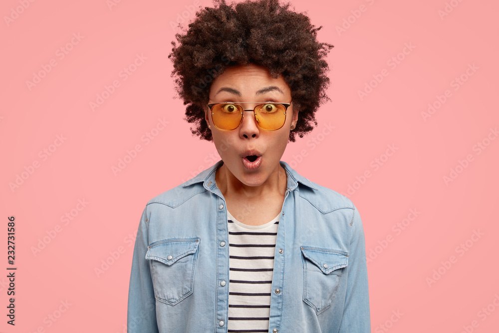 Shocked stupefied emotional young African American woman has frightened and surprised look, opens mouth widely, wears yellow sunglasses, denim shirt, stares at camera, isolated over pink background