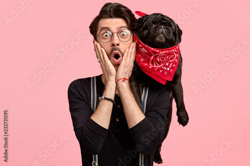 Emotive unshaven young man keeps both hands on cheeks, has his lovely black dog on shoulders, cant believe in shocking news, isolated over pink background. People, animals and reaction concept