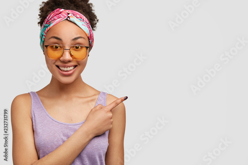 Glad pretty black female model points with index finger aside, wears yellow shades, casual clothing, shows free space for your advertisement or promotional content. Ethnic girl promots item.