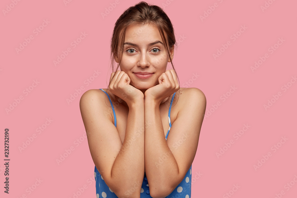 Charming beautiful female model keeps both hands under chin, looks seriously directly at camera, wears casual dress, has healthy skin, models against pink studio background. People and beauty concept Charming beautiful female model keeps both hands under chin, looks seriously directly at camera, wears casual dress, has healthy skin, models against pink studio background. People and beauty concept