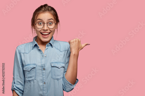 Indoor shot of hilarious content woman smiles broadly, poses aside with thumb, being glad to advertise some object, isolated over pink backgrund, shows space for your advertisement or promotion