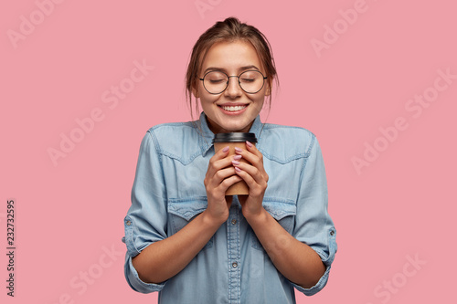 Pleased attractive woman holds takeaway coffee, smiles broadly at camera, warms herself with hot beverage, wears casual shirt, isolated over pink background. People, drink and lifestyle concept