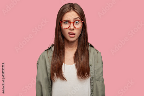 Fototapeta Indoor shot of indignant woman frowns face in displeasure, wears spectacles, shirt, models against pink background, doesnt like something dressed in casual shirt