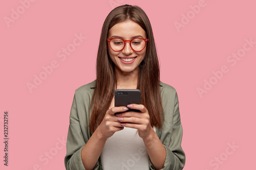 Satisfied lovely woman holds modern cell phone, texts with friend online in chat, dressed in stylish clothes, poses against pink wall, connected to high speed internet. Shot of teenager with device