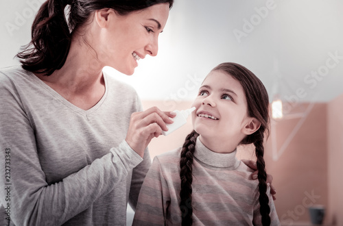 Caring about health. Portrait of smiling mother giving nasal drops to her daughter while the daughter smiling at her mother
