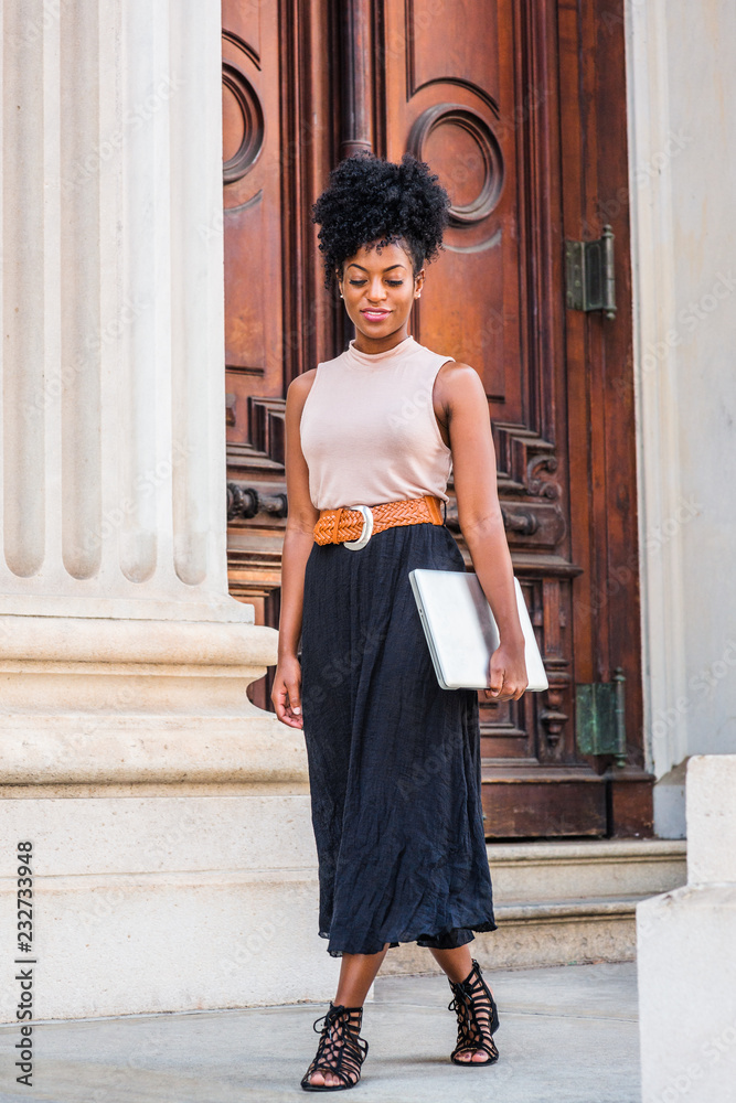 Young black woman with afro hairstyle, wearing sleeveless light color top, black skirt, belt, strappy sandals, carrying laptop computer, walking by office doorway in New York, looking down, thinking..