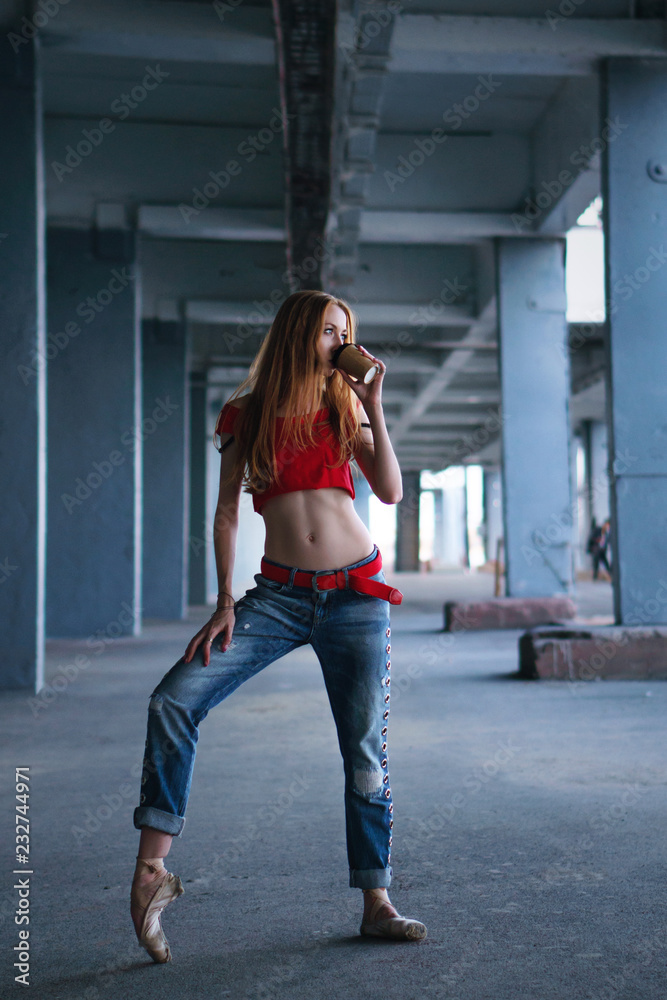 Ballerina dancing with a cup of coffee in jeans, t-shirt and pointe. Street performance. Modern ballet. Slender girl holding a paper cup with a hot drink.