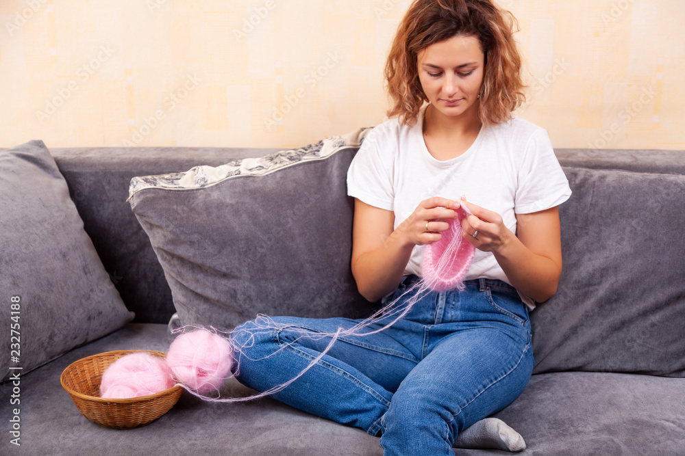 Fototapeta premium Close up of a young married woman dressed in trendy d jeans and t-shirt knitting a pink hat with knitting needles in home. Freelance creative working and living concept