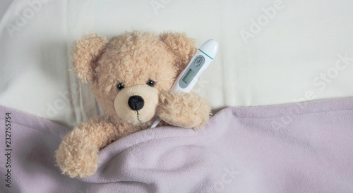 Cold, flu or allergy. Cute teddy in bed, covered with a warm blanket, holding a tissue