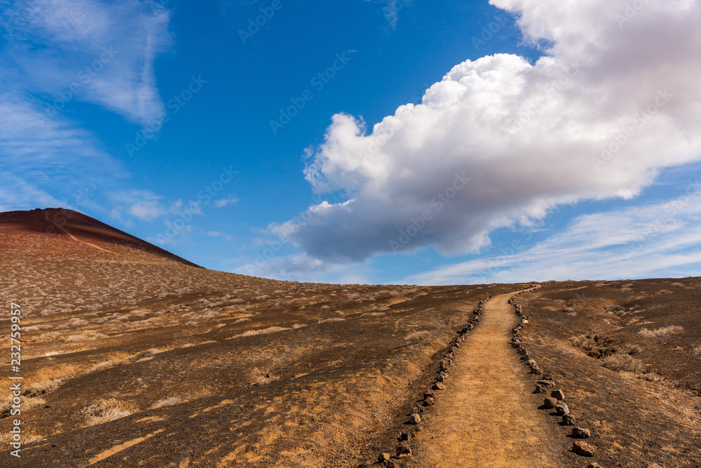 Dirt trail going through a spectacular volcanic landscape towards a red rock mountain on a blue sky summer day. Hiking path in La Graciosa Island, Canary, Spain.