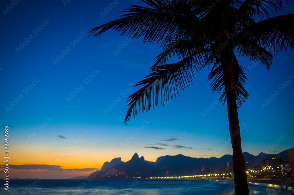 Scenic sunset view of Ipanema Beach with silhouette of a palm tree in front of the dramatic city skyline of Rio de Janeiro, Brazil