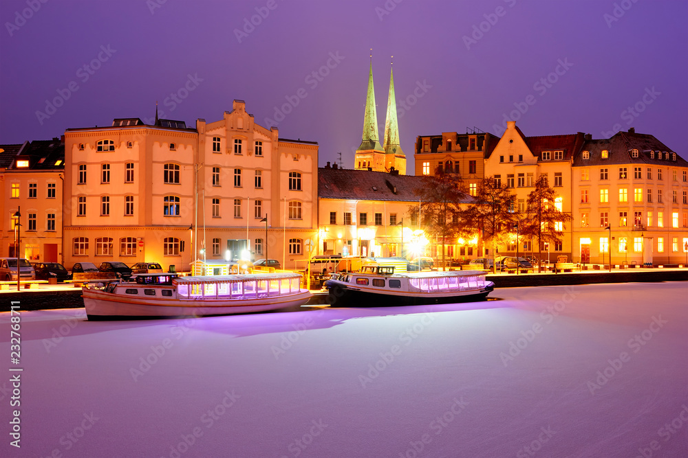 Boats on Frozen Trave river at nights. Lights on Lubeck city emb